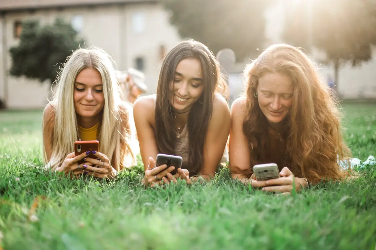three teenagers laying in grass using iphones.